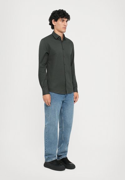 Cămașă cu mâneci lungi, de culoare verde închis, purtată împreună cu blugi din denim ușor albastri și adidași negri. Design simplu, material ușor, stil ajustat.