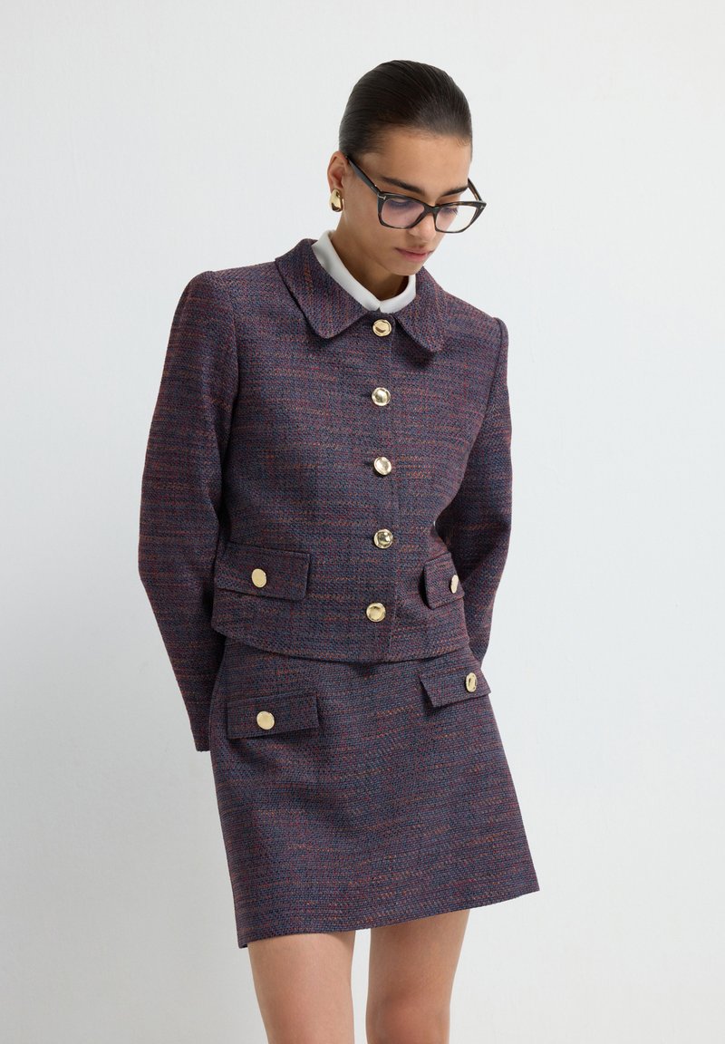 Giacca di tweed in viola scuro con dettagli rossi e bottoni dorati, abbinata a una gonna coordinata. Presenta un ampio colletto e due tasche frontali.