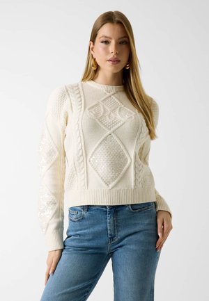 Frau mit einem cremefarbenen, kabelgestrickten Pullover mit Pailletten-Diamantmustern, kombiniert mit blauen Jeans, stehend vor einem weißen Hintergrund.
