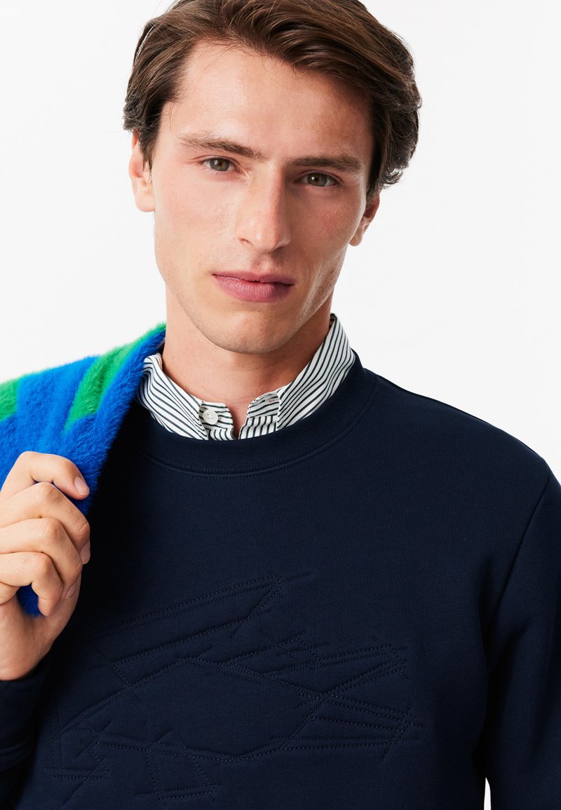 Granatowy sweter z teksturowanym wzorem, z okrągłym dekoltem i ściągaczami, noszony na niebieskiej i zielonej koszuli w paski z kołnierzykiem.