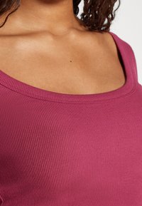Detailní záběr osoby v úzkém, tmavě růžovém topu s výstřihem, který odhaluje klíční kost a část kudrnatých vlasů.