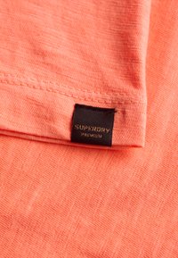 Camicia arancione corallo con tessuto strutturato e motivo sottile. Etichetta nera con scritta "SUPERDRY PREMIUM" in lettere dorate.