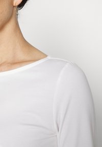 Närbild av en person som bär en vit långärmad skjorta och en tunn gyllene hoopörhänge mot en enkel ljus bakgrund.