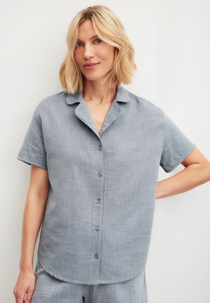 Femme aux cheveux blonds portant une chemise gris clair à manches courtes et boutonnée, assortie à un short, debout avec une main sur la hanche.
