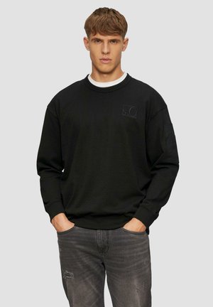 Zwarte sweatshirt gemaakt van stof met ribbels aan de cuffs en de zoom, met een subtiele logo-detail op de borst, gecombineerd met versleten grijze jeans.