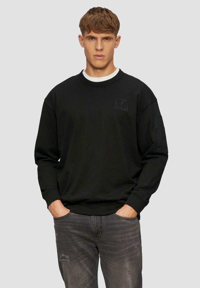 Zwarte sweatshirt gemaakt van stof met ribbels aan de cuffs en de zoom, met een subtiele logo-detail op de borst, gecombineerd met versleten grijze jeans.