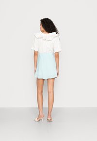 Blusa branca com decote de babados, combinada com uma saia mini texturada de azul claro. Estilizada com saltos pálidos, vista pela parte de trás.