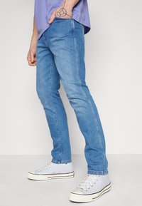 Man som bär ljusblå slim-fit jeans, vita hög-top sneakers och en lila skjorta, står med ena handen i fickan.