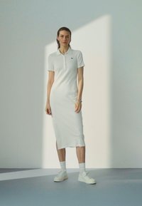 Biele polo šaty s krátkymi rukávmi, slim fit, rebrovaná textúra, po kolená, so malým logom na hrudi, v kombinácii s bielymi teniskami.