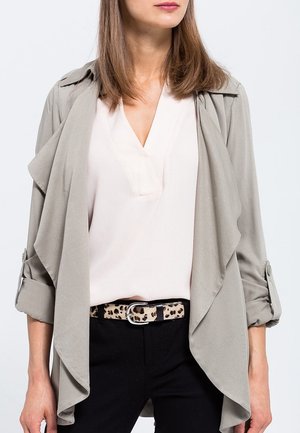 Femme portant une veste drapée beige clair, un chemisier blanc à col en V, un pantalon noir et une ceinture imprimée léopard, vue du cou aux hanches.