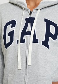Primer plano de una sudadera con cremallera gris con cordones blancos y grandes letras azules marino "GAP" cosidas en el pecho.