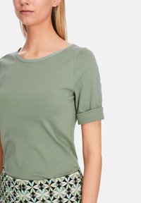Leicht tailliertes grünes T-Shirt mit rundem Ausschnitt und umgeschlagenen kurzen Ärmeln. Glatter Stoff mit subtilen Nähdetails.