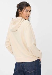 Beige hoodie met een trekkoordkap, zachte stof, geribde manchetten en een losvallende pasvorm, gecombineerd met spijkerbroeken. Achteraanzicht weergegeven.