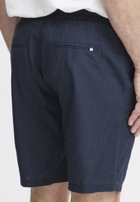 Tamsiai mėlynos lino šortai su elastingu liemeniu ir dviem užpakalinėmis kišenėmis. Ant užpakalinės kišenės yra mažas baltas akcentas. Minkšta tekstūra.