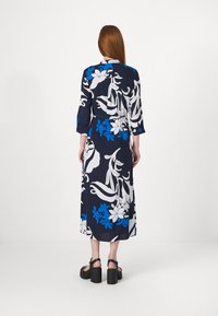 Vestido floral azul-marinho com padrões brancos e azuis, mangas de três quartos e cintura cingida por um cinto, combinado com sandálias pretas de plataforma.