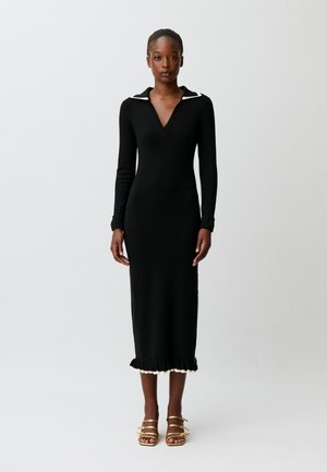 Donna che indossa un lungo vestito nero con colletto bianco e dettagli sull'orlo, in piedi su uno sfondo bianco semplice, con tacchi dorati con cinturini.