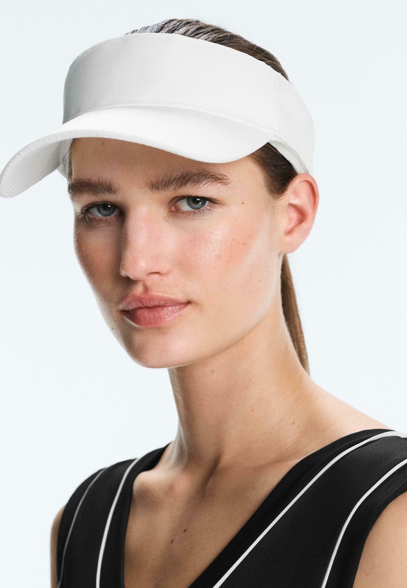 Jeune femme à la peau claire et aux yeux bleus portant une visière blanche et un haut noir sans manches avec un bord blanc, regardant légèrement sur le côté.