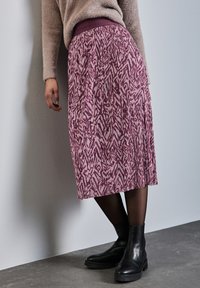 Gonna midi viola con un motivo, dotata di una vita elastica testurizzata, abbinata a stivali neri alla caviglia e collant neri trasparenti su un pavimento grigio.