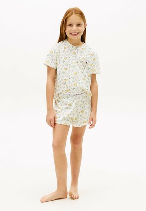 Lachend meisje met lang rood haar, gekleed in een wit kort mouwbloesje met bloemenprint en bijpassende korte broek met het Tommy Hilfiger-logo op de tailleband, staand op blote voeten.