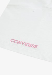 Balta medvilnės marškinėliai su rožiniu "CONVERSE" logotipu, atspausdintu netoli apatinio krašto, matoma siuvimo linija ir lygi tekstūra.