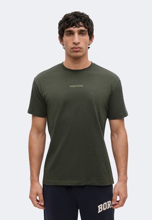 Man met kort krullend haar, die een donkergroen Björn Borg T-shirt en marineblauwe shorts draagt, staand tegen een effen lichte achtergrond.