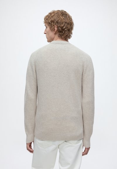 Homme aux cheveux blonds bouclés portant un pull beige et un pantalon blanc, debout de dos sur un fond blanc uni.