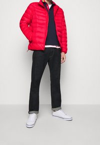 Röd puffjacka med dragkedja framtill, med ståkrage, bärs över en marinblå polo och mörka jeans, tillsammans med vita sneakers.