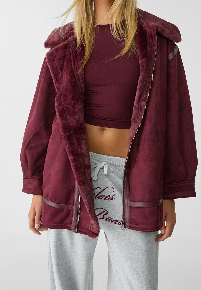 Femme portant une veste bordeaux doublée de fausse fourrure sur un crop top assorti et un pantalon de survêtement gris clair avec des lettres scriptes bordeaux.