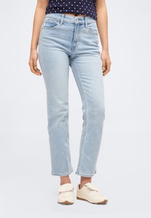 Žena nosí světle modré džíny se širokými nohavicemi, tmavě modrý top s puntíky a bílé tenisky na jednobarevném pozadí.