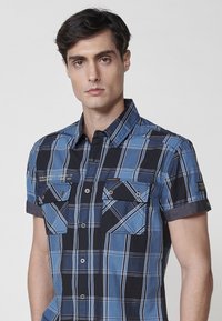 Korte mouwen geruite shirt in blauw, zwart en wit. Heeft een button-down kraag, twee borstzakken met kleppen en contrasterende opgerolde mouwen.