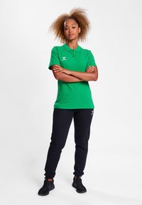Grön polo skjorta med krage och logotyp, kombinerad med svarta slim-fit byxor och svarta sportskor. Slät textur, avslappnad design.