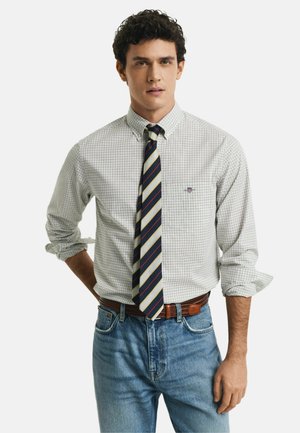 Camisa de cuadros blancos con mangas arremangadas, combinada con una corbata a rayas en azul, amarillo y rojo. Llevada con jeans de mezclilla azul claro y cinturón marrón.
