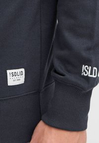Dunkelblaues Sweatshirt aus weichem Material, mit gerippten Bündchen, Seitennahtdetails und einem weißen Logoetikett mit dem Druck "!SOLID DENMARK EST.1999".
