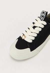 Svart lærersneaker med teksturerte svarte aksenter, kremfargede lisser og gummisåle. Har et unikt mønster på hælen og metalldetaljering.