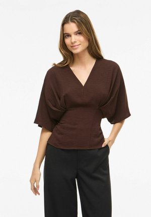 Jeune femme portant un haut marron foncé à col en V avec de larges manches et un pantalon noir, debout avec une main dans la poche sur fond blanc.