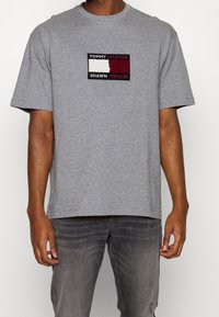 Harmaa puuvillapaita lyhyillä hihoilla, jossa on edessä suorakulmainen brodeerattu logo punaisena, valkoisena ja mustana.