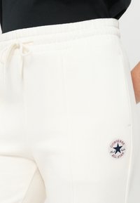 Hvite joggebukser i fleece med elastisk midjebånd, frontlommer og en rund blå-og-rød "Converse All Star"-logo på låret.