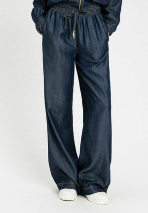 Persona che indossa pantaloni denim blu scuro a gamba larga con vita elastica e coulisse, mani nelle tasche, abbinati a sneaker bianche.