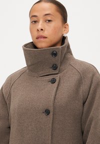 Brūns vilnas mētelis ar augstu apkakli, četriem melniem pogām un teksturētu apdari. Dizainā ir iekļautas raglāna piedurknes un pieguļoša siluets.