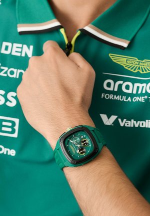 Zaļš kvarca pulkstenis ar kvadrātveida korpusu, melniem akcentiem un hronogrāfa funkcijām. Nēsāts uz rokas, kopā ar zaļu polo krekli.