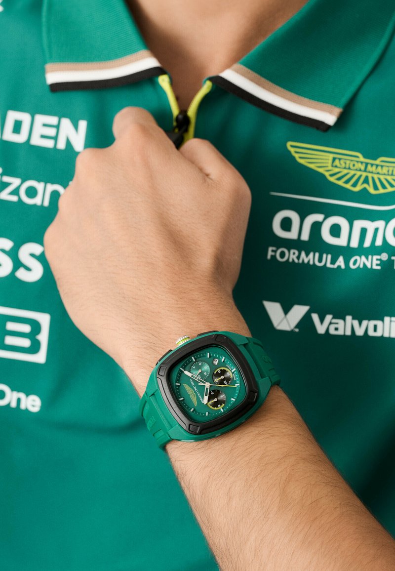 Zaļš kvarca pulkstenis ar kvadrātveida korpusu, melniem akcentiem un hronogrāfa funkcijām. Nēsāts uz rokas, kopā ar zaļu polo krekli.