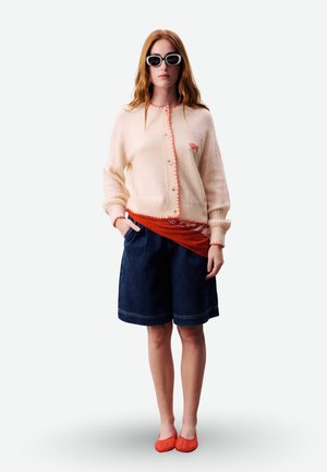 Femme aux longs cheveux roux portant des lunettes de soleil noires, un cardigan crème, une chemise orange, un short en jean foncé et des ballerines orange, debout devant un fond blanc.