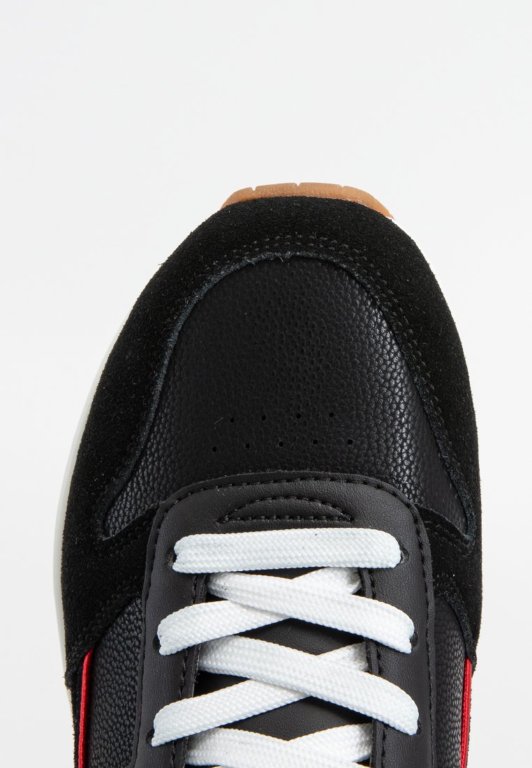Zwarte sneaker met een getextureerde leren neus en suède panelen, witte veters en een klein gedeelte van de bruine rubberen zool zichtbaar aan de bovenkant.