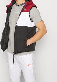 Mężczyzna w bezrękawniku puchowym w kolorach czerwonym, białym i czarnym, ubrany w szary t-shirt i białe spodnie, stoi z jedną ręką w kieszeni kamizelki.