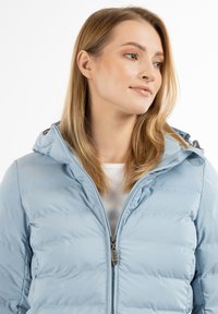 Chaqueta tipo puff en azul claro con cierre frontal de cremallera, capucha y textura acanalada; cuenta con un ligero brillo y un logo en el tirador de la cremallera.