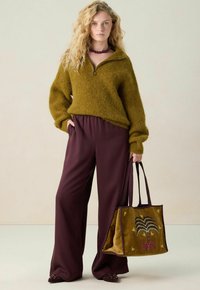 Eine Frau steht mit einem olivgrünen, flauschigen Zip-Pullover und weiten bordeauxroten Hosen da und hält eine passende olivgrüne Tragetasche mit besticktem Design.