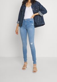 Jeans de mezclilla de lavado claro con cintura alta y rasgaduras visibles; combinados con una blusa blanca y una chaqueta de mezclilla oscura. Lleva sandalias blancas con tiras.