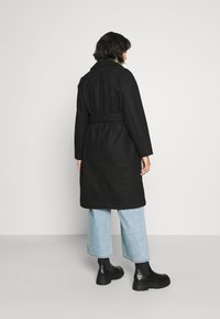 Black belted coat with a tailored design and large collar, featuring a smooth texture. Worn over light blue wide-leg jeans and black boots.