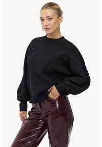 Sweat-shirt noir surdimensionné avec un col rond et des manches larges, associé à un pantalon bordeaux à forte brillance. Une bague proéminente portée à la main.