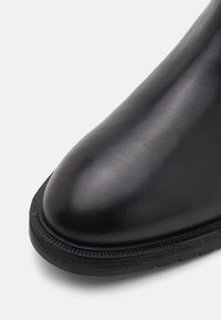 Svarte skinnankelstøvler med en glatt, blank finish. Rund tå, teksturert gummisåle og subtile sømdetaljer langs kantene.